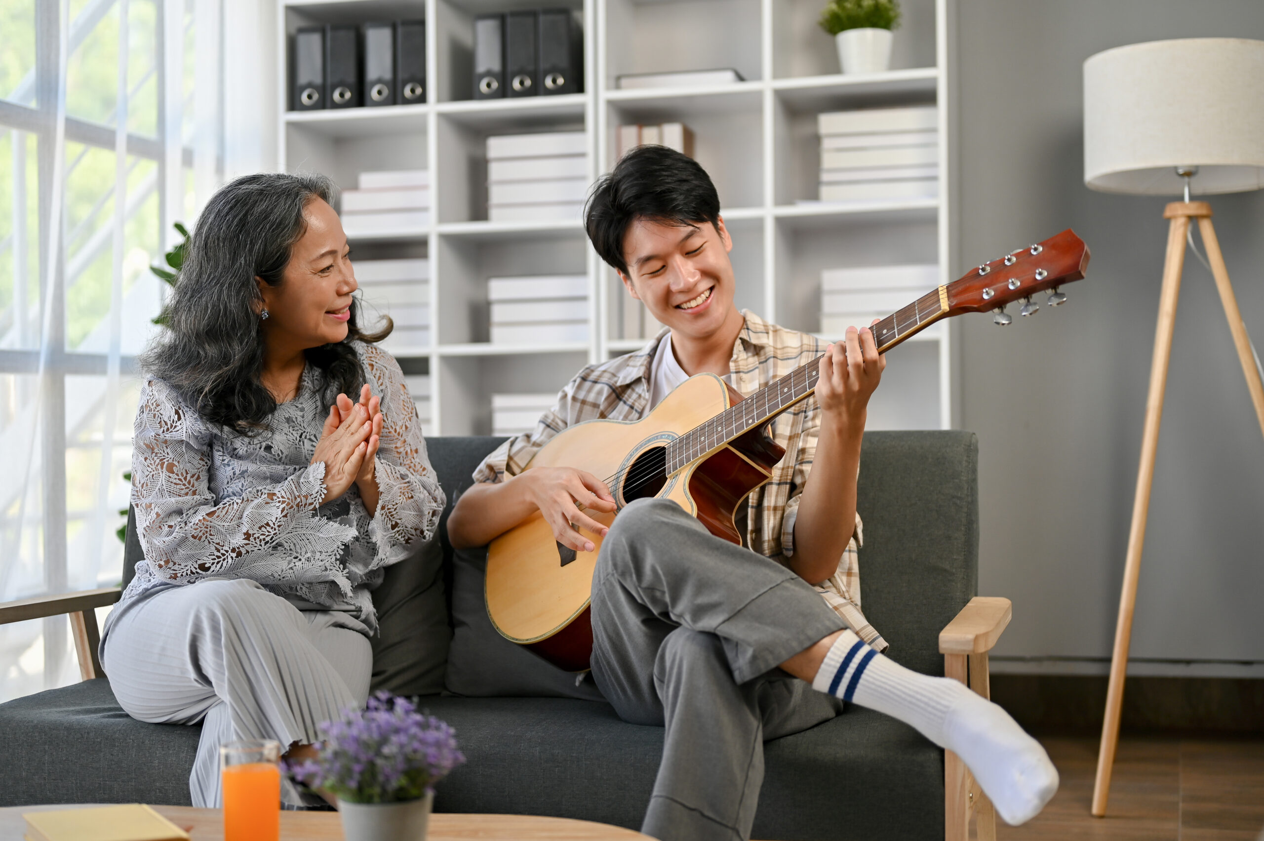 A happy Asian grandmother is clasping her hands while listening to her grandson sing and play guitar at home together.